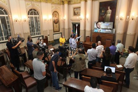 vários repórteres e cinegrafistas em sala assistindo ao prefeito falar. Ele aponta para um telão na frente da sala onde há dados sobre a covid-19. #paratodosverem