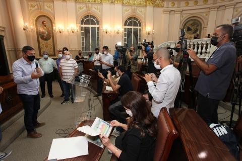 o prefeito está em pé segurando um microfone. Ele está dando entrevista em uma sala com repórter, cinegrafistas e outras pessoas. #paratodosverem