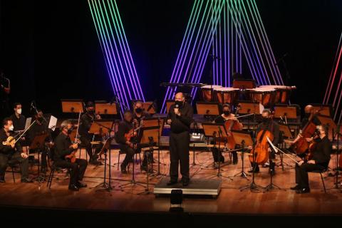 maestro está no palco de costas para os músicos e de frente para o público. Ele fala ao microfone. #paratodosverem