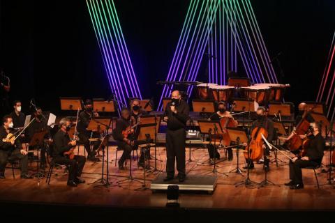 maestro está no palco de costas para os músicos e de frente para o público. Ele fala ao microfone. #paratodosverem