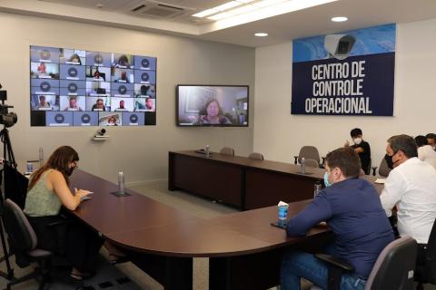 pessoas reunidas em torno de uma mesa em formato de U. Ao fundo, um monitor de computador e uma imagem reproduzida num telão, com os demais participantes on-line. #paratodosverem