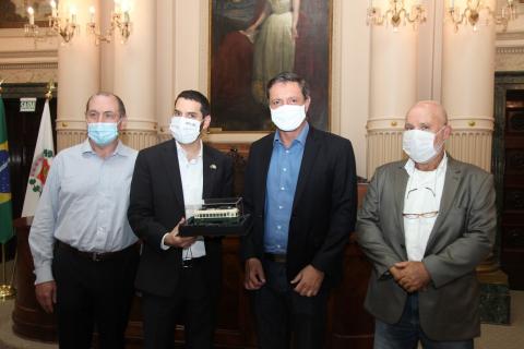 Consul segura réplica do bonde posando para foto com o prefeito e mais duas pessoas. #Paratodosverem