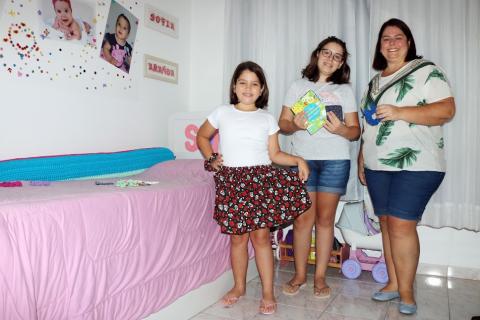 Alessandra posa para foto ao lado de duas filhas. A mais nova, ao lado de uma cama, segura um shorts que a mãe costurou. #paratodosverem