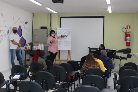 Aula teórica. Cadeiras com pessoas sentadas e uma pessoa em pé à frente de um telão. #paratodosverem