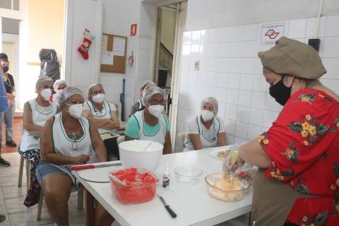 alunas observam preparo de massa por uma mulher atrás de mesa. #paratodosverem