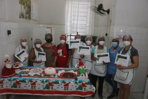 Alunas posam para foto atrás de mesa exibindo seus certificados de participação. #paratodosverem