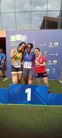 Três atletas em pé, abraçadas, mostram medalhas no pódio. #pratodosverem