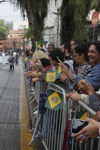 Pessoas com bandeiras do Brasil próximas ao gradil do desfile. #paratodosverem
