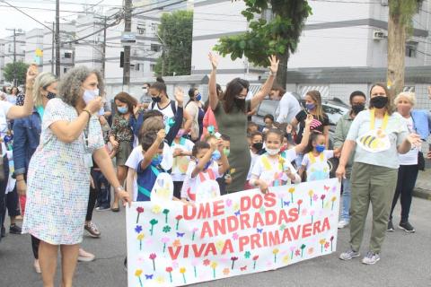 crianças caminham pela rua carregando faixa. O grupo saúda as pessoas na rua erguendo os braços. #paratodosverem