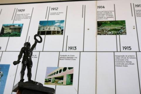 Painel com linha do tempo do esporte santista e um troféu à frente. #paratodosverem