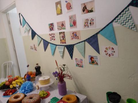 parede decorada com bandeirinhas e imagens coloridas. Mesa com bolos, frutas e flores abaixo. #paratodosverem 