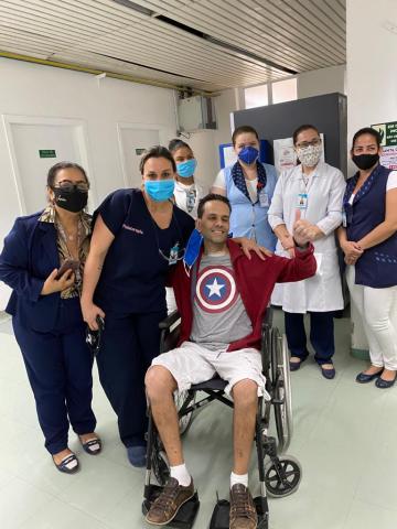 Marco, sentado em cadeira de roda, ao lado de profissionais de saúde no dia da alta do hospital. #pracegover