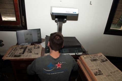 homem está sentado diante de aparelho de scanner e tela de computador. Tanto do lado esquerdo quanto do direito há pilhas de jornais antigos. #paratodosveremaratodosverem