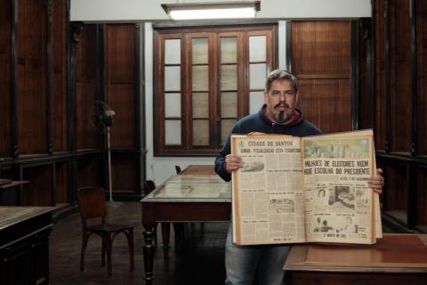 Homem segura caderno com várias edições do extinto jornal Cidade de Santos. Ele está em pé segurando o caderno aberto em uma página. #paratodosverem