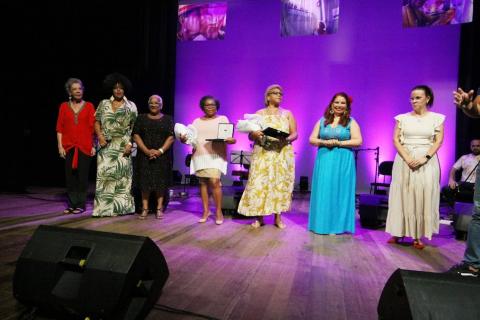 Sete mulheres estão no palco. Duas delas seguram ramalhetes de flores. #paratodosverem