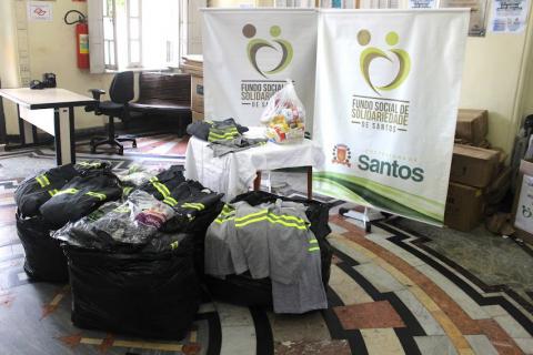 vários sacos plásticos com roupas em cima estão no chão. ao fundo, dois banners do fundo social de solidariedade. #paratodosverem 