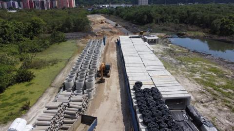 vista geral da área onde estão sendo realizadas as obras, com tubos e aduelas empilhados, e vegetação ao lado. #paratodosverem