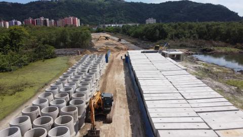 tubos e aduelas em terreno com tratores. Nas laterais, vegetação. E, ao fundo, prédios do bairro do saboó. #paratodosverem