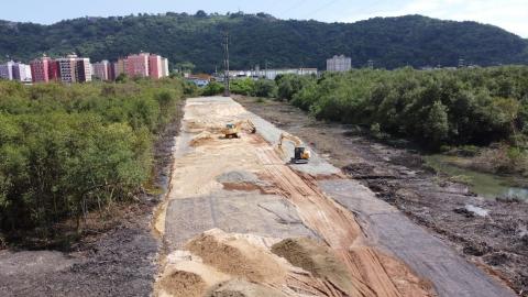 grande área coberta de terra onde será realizada a obra, ladeada por vegetação e prédios ao fundo. #paratodosverem