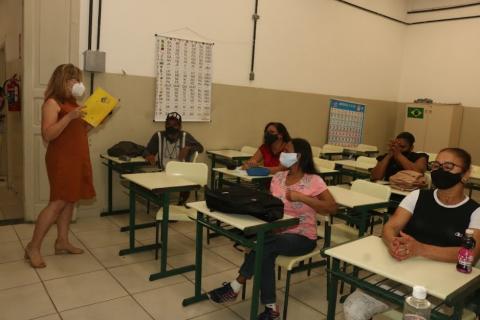 Em sala de aula, professora lê para alunas sentadas. #paratodosverem