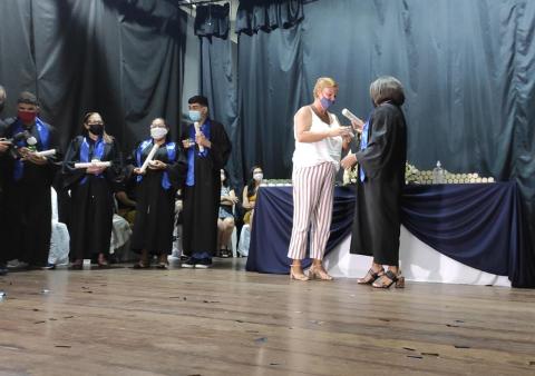 mulher entrega canudo com diploma a aluna que veste beca.Elas estão no palco. Há outros alunos no mesmo lugar. #paratodosverem