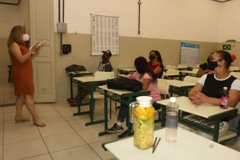 sobre a mesa há uma garrafa com água e rodelas de limão. A professora está em pé e os alunos, sentados. #paratodosverem