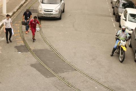 homem faz embaixadinha nomeio da rua, passando entre os tilhos do bonde. Há carros e uma moto da CET acompanhando. #paratodosverem