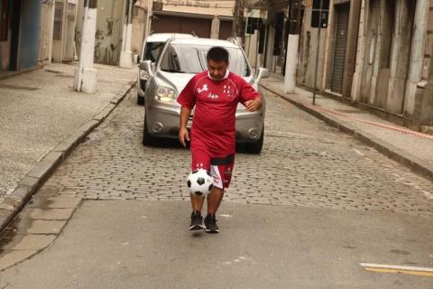HOmem faz embaixadinha no meio da rua. Ele usa uniforme de jogador de futebol e chuteiras. #paratodosverem