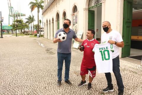 Ricardinho está entre o prefeito, que segura uma bola, e o secretário de esportes, que segura uma camisa com o número 10 e o nome de Pelé nas costas. Ao fundo, a fachada do Museu Pelé. #paratodosverem