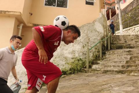 homem equilibra bola no pescoço, entre a cabeça e as costas. Ele está subindo escadarias de morro. Ouro homem acompanha. #paratodosverem