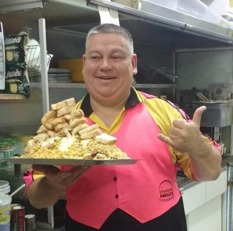 Paulo Roberto Almeida de Sá segura um prato cheio de comida, com o quiosque ao fundo. #paratodosverem