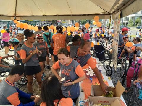 várias pessoas reunidas sob tenda. Ao fundo, balões cor de laranja. #paratodosverem