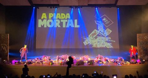 dançarinos estão no palco em coreografia. A grande maioria está deitada. Dois estão em pé. Ao fundo, em um painel iluminado se lê Piada Mortal