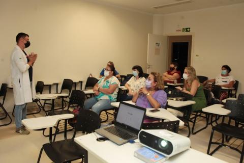 homem está em pé à esquerda falando a várias pessoas que estão sentadas em carteiras estudantis. Em primeiro plano, um notebook aberto. #paratodosverem