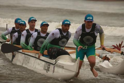 Equipe Dos Reis Va’a chega na beira da água. O líder do grupo sai correndo da canoa. Um homem atrás dele começa também a sair. Outros quatro estão sentados na canoa. #paratodosverem