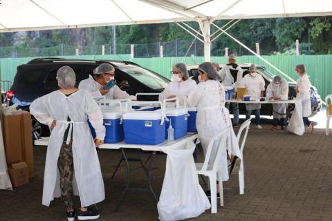 esquema de vacinação montado em drive-thru. Mulheres vestidas de branco estão pé ao lado de mesas onde estão dispostos coolers para as vacinas. #paratodosverem