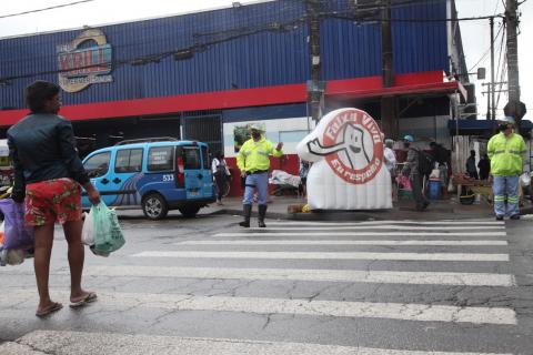 agentes da CET estão de um lado da rua ao lado de inflável da campanha. Mulher, do outro lado, inicia a travessia. #paratodosverem