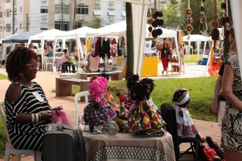 Mesa com bonecas afro e mulher sentada ao lado #paratodosverem