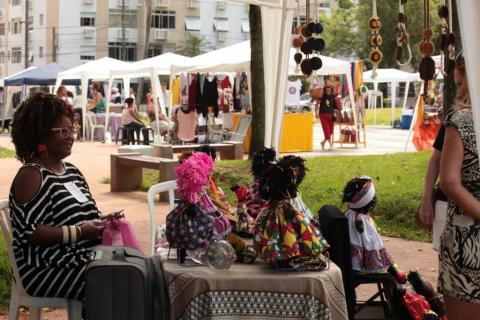 Mulher sentada e mesa com artesanatos afro #paratodosverem