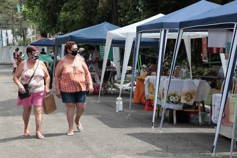 Mulheres observam barraca com toalhas de mesa. #pratodosverem