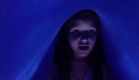 menina está no escuro, com parte de uma coberta cobrindo sua cabeça. Ambiente é iluminado ´por uma luz azul. #paratodosverem
