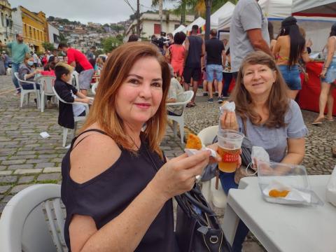 Duas mulheres sentadas em mesa comem salgado e bebem cerveja #paratodosverem