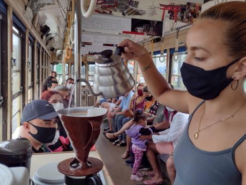 Mulher prepara café dentro do bonde temático com passageiros. #pracegover