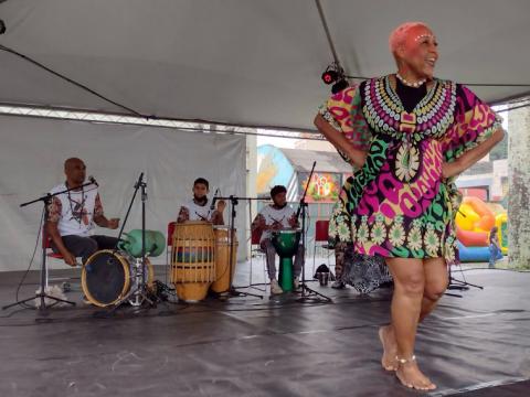 Mulher se apresenta com dança africana com músicos ao fundo. #pracegover