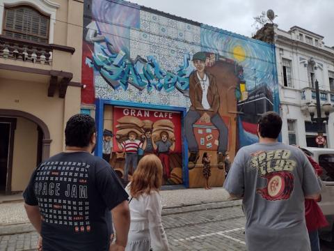 Dois homens e uma mulher, de costas, observam o Mural do Imigrante na Rua do Comércio. #pracegover