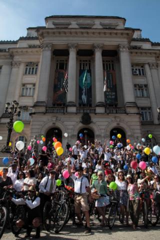 Dezenas de ciclistas, muitos trajados com roupas de época, posam para foto em frente à Prefeitura. #pratodosverem
