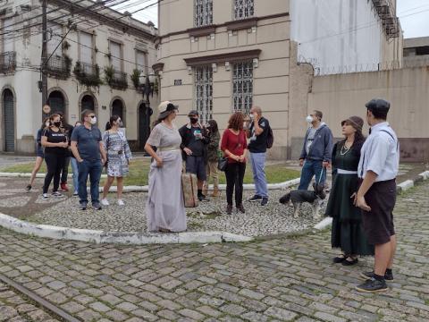 Pessoas participam de passeio a pé com atores vestidos de roupas de época. #pracegover