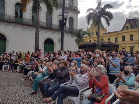 Pessoas assistem apresentação e aplaudem sentadas e em pé. #pracegover 