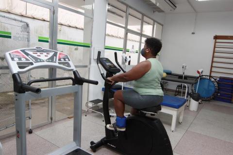 mulher está sentada em cadeira ergométrica pedalando. Há outros equipamentos na sala. #paratodosverem