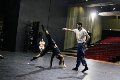 bailarina está com as mãos no solo e as pernas para o alto. Orientador está em pé acompanhando o movimento. #paratodosverem